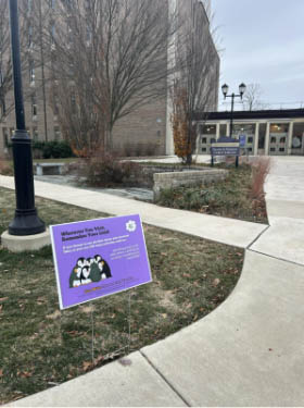 A purple campus sign on a lawn near a sidewalk and academic building. The sign features cartoon penguins and promotes responsible alcohol choices as part of a “Live Well: Choose Before You Use” campaign.