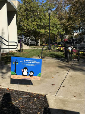 Sidewalk near a campus building entrance with a small yard sign featuring cartoon penguins and a message, surrounded by grass, trees, and a bike rack.
