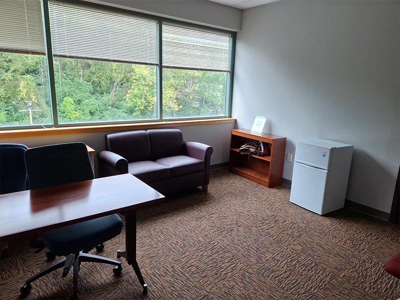 From the entry, a large room with windows that have moveable blinds, a mini-fridge, a couch, and a table with office chairs. 