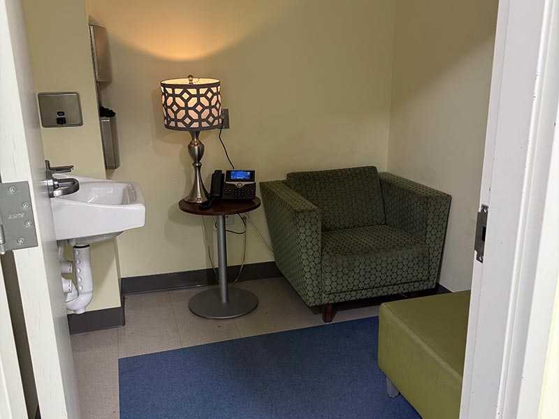  From the entry, a small room with a sink, mirror, soap and paper towel dispenser on the right wall. On the back wall, an armchair with a small table with a lamp and a phone. In front of the armchair, a soft ottoman. A blue rug covers the floor.  