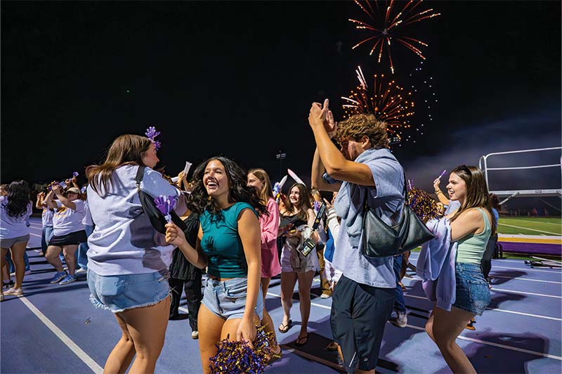 A group of students chatting and laughing at Ram Rally under the Lights while fireworks are going off in the background