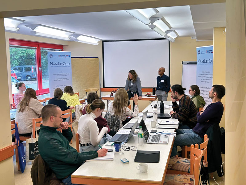 Around a dozen students with laptops sitting at a long table in a classroom. Teachers at the front with a blank white board.