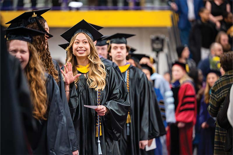 A smiling graduate in a cap and gown waves while walking in a graduation ceremony procession.