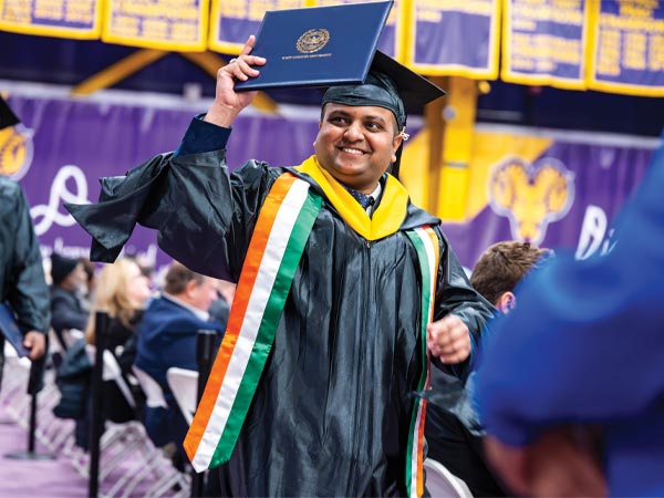 A Graduate student at commencement wearing their cap and gown excitedely holds their diploma folder up.