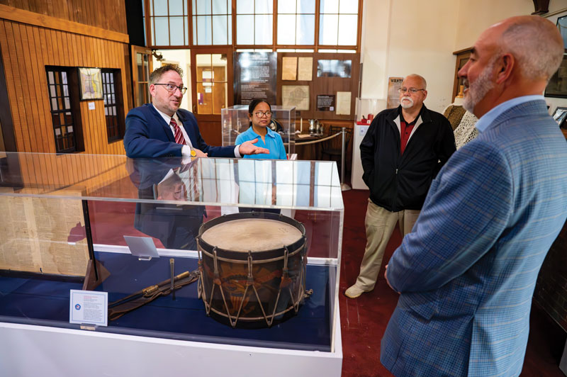 4 people stand and chat over a glass case containing items, including a drum