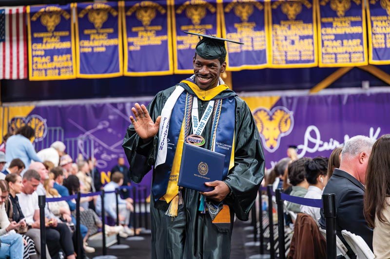 A student in graduation robes waves and holds his diploma