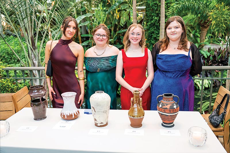 4 women dressed up in their gowns smile behind a table with vases on it