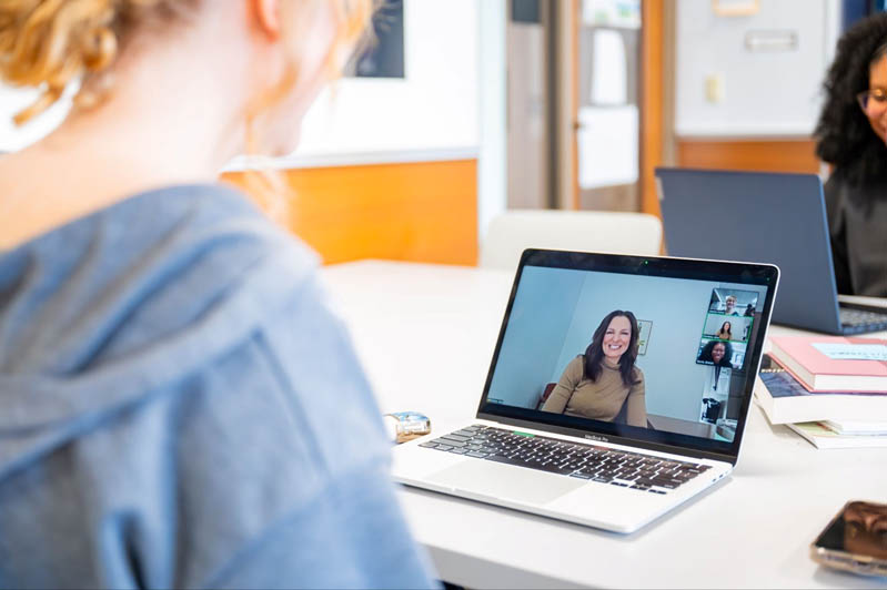 large image. A person sits at a table using a laptop for a video call. On the screen, a woman smiles and waves, with smaller participant windows visible, indicating a group meeting. Another person is seated in the background, and books and a phone rest on the table.