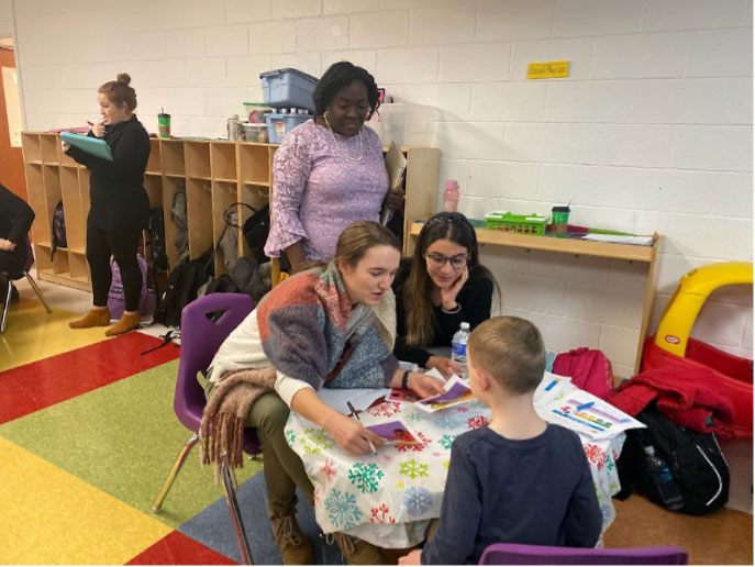 Classroom setting. 2 women sitting at a circular table helping a child.