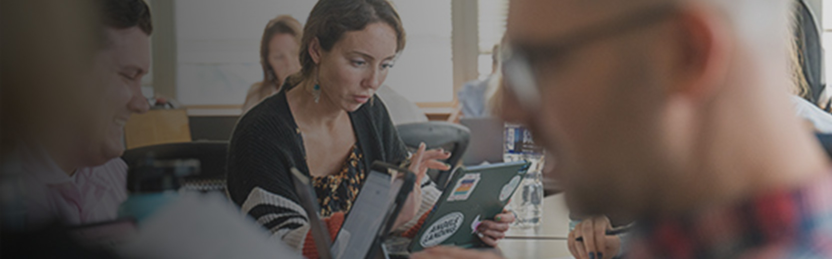 A group of adults seated at a table in a classroom or meeting setting, focused on their laptops and tablets, with one woman in the center looking intently at her screen while others work around her.